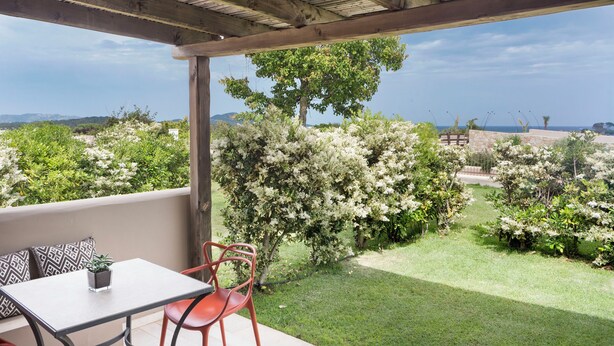 A covered patio with a table and chairs, a view of a landscaped garden with flowering bushes, and a distant landscape.