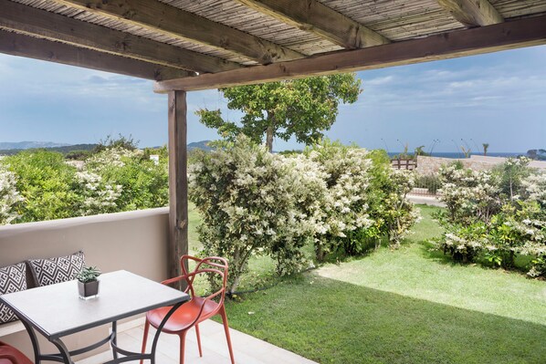 A covered patio with a table and chairs, a view of a landscaped garden with flowering bushes, and a distant landscape. - Baglioni Resort Sardinia (San Teodoro)