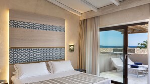 Habitación de hotel con cama, mesita de noche, cabecera estampada, ventana grande con vista al mar y balcón con una silla y una mesa.