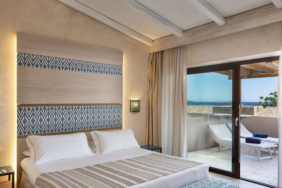 Une chambre d’hôtel avec un lit, une table de chevet, une tête de lit à motifs et une porte-fenêtre coulissante donnant sur un balcon avec vue sur la mer.