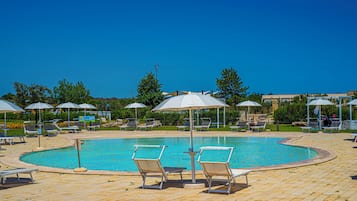 Ein Schwimmbecken mit Liegestühlen und Sonnenschirmen, umgeben von Grünpflanzen und blauem Himmel.