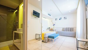 A modern hotel room with a glass shower enclosure, a bed with a blue accent, a bedside table, and a small desk with a chair. - Relais Masseria Le Cesine Cdshotels (Vernole)