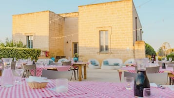An outdoor dining area with tables set for guests, a stone building in the background, and string lights overhead.