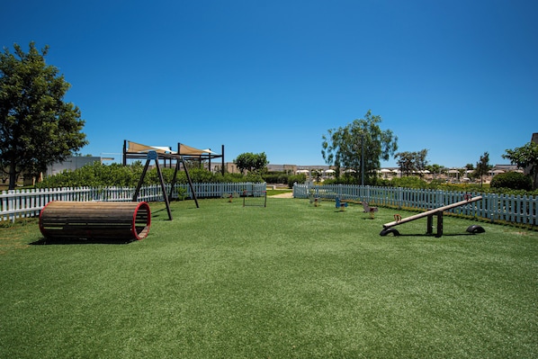 Un parque infantil con diversos juegos, una cerca blanca y un edificio al fondo.