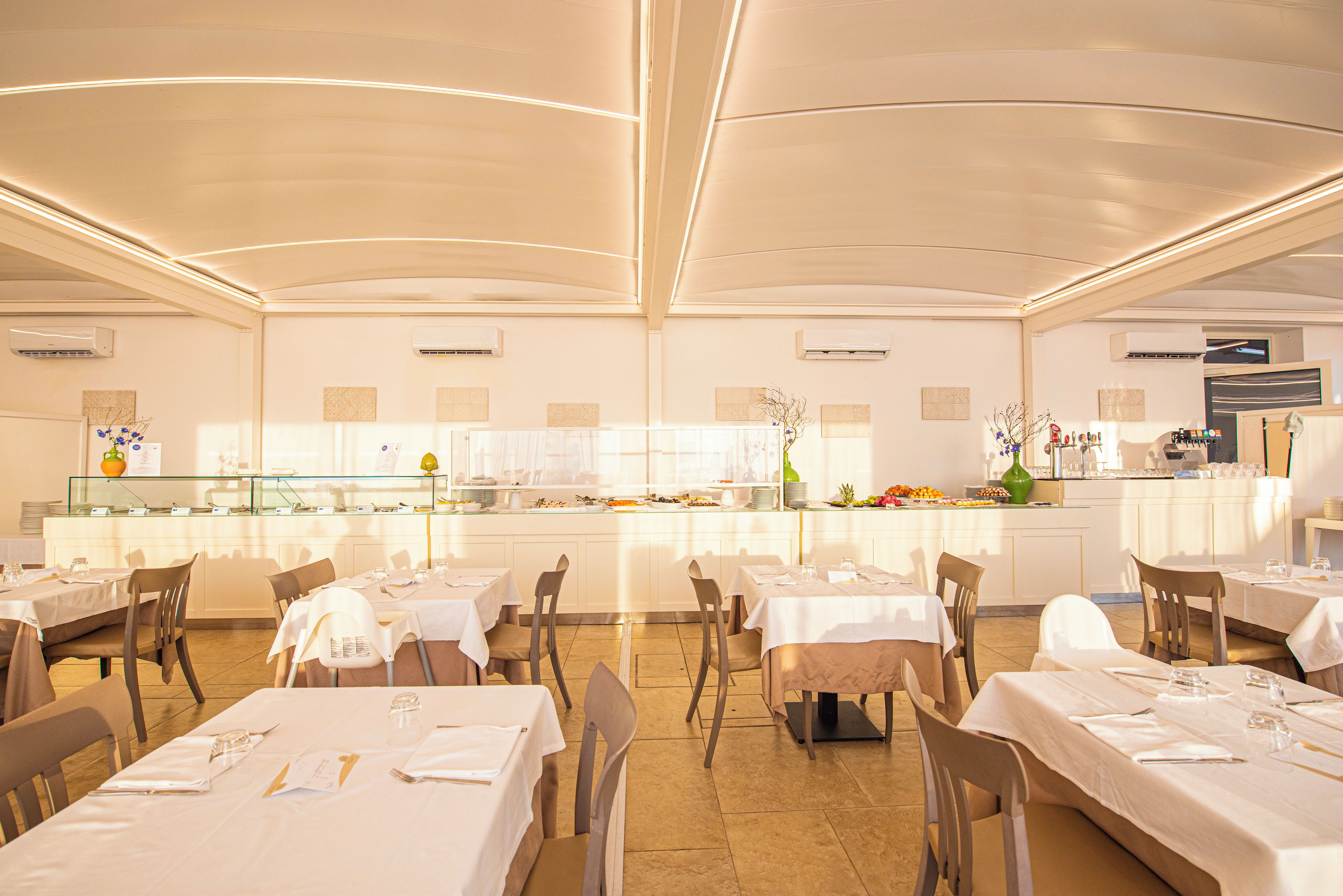 A spacious restaurant with multiple tables and chairs, a central counter with a display of food items, and a modern ceiling design.