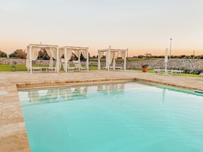 Una piscina con superficie de agua azul cristalina, rodeada de baldosas de piedra. Hay toldos blancos con cortinas que ofrecen sombra sobre sillones de descanso.