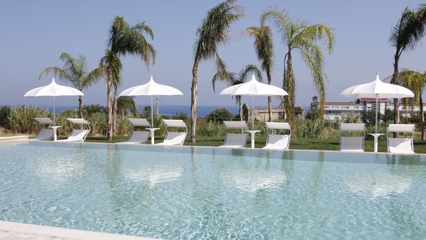 A pool with clear water, surrounded by white lounge chairs and umbrellas, palm trees, and a view of the ocean. - Il Borghetto Creative Resort (Tropea)