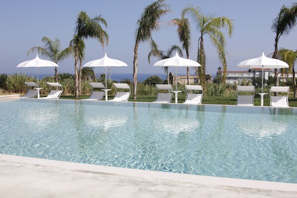 A pool with clear water, surrounded by white lounge chairs and umbrellas, palm trees, and a view of the ocean. - Il Borghetto Creative Resort (Tropea)