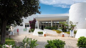 Un bâtiment moderne à l’extérieur blanc, doté d’une entrée abritée et d’une cour pavée agrémentée de plantes en pot et d’un arbre.