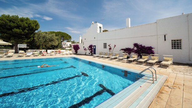 Une piscine à l’eau d’un bleu cristallin, entourée de chaises longues et bordée d’un bâtiment blanc doté d’un toit en tuiles.