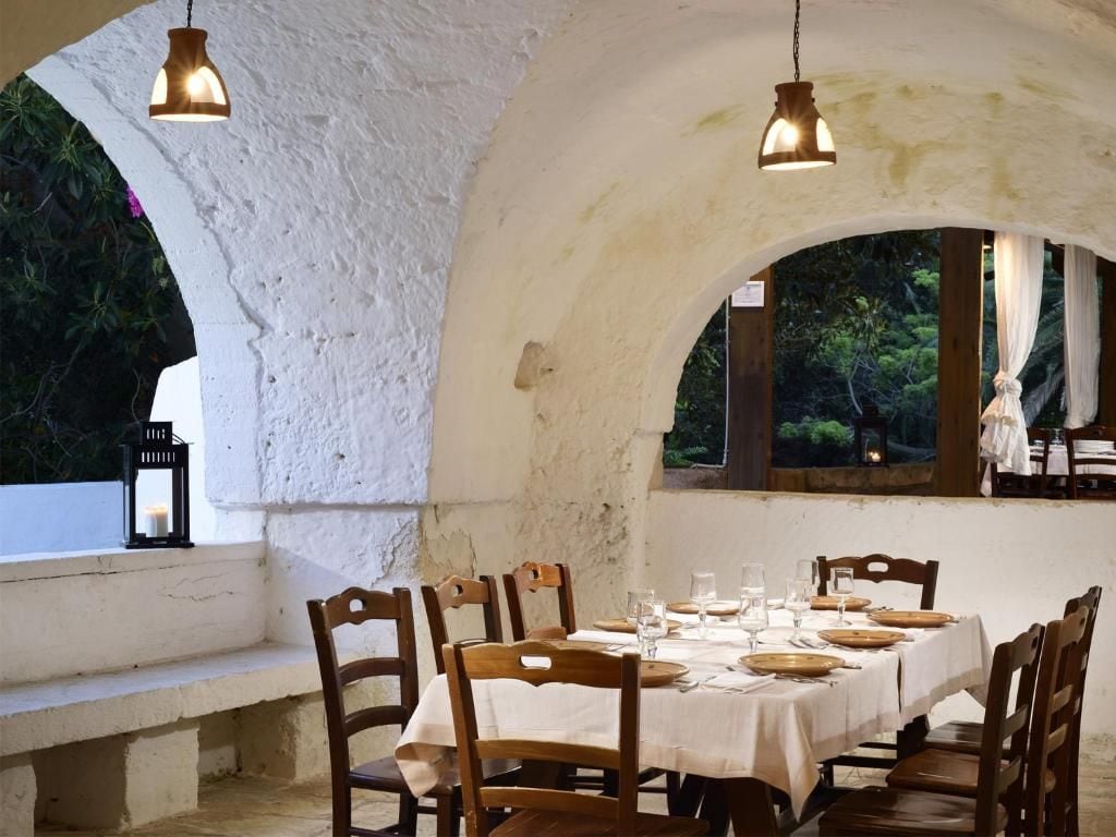 Un'area pranzo all'aperto con un tavolo bianco apparecchiato per sei persone, sedie di legno e lampade a sospensione.