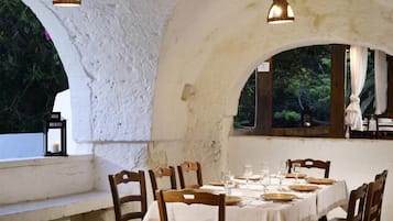 Un'area pranzo all'aperto con un tavolo bianco apparecchiato per sei persone, sedie di legno e lampade a sospensione.
