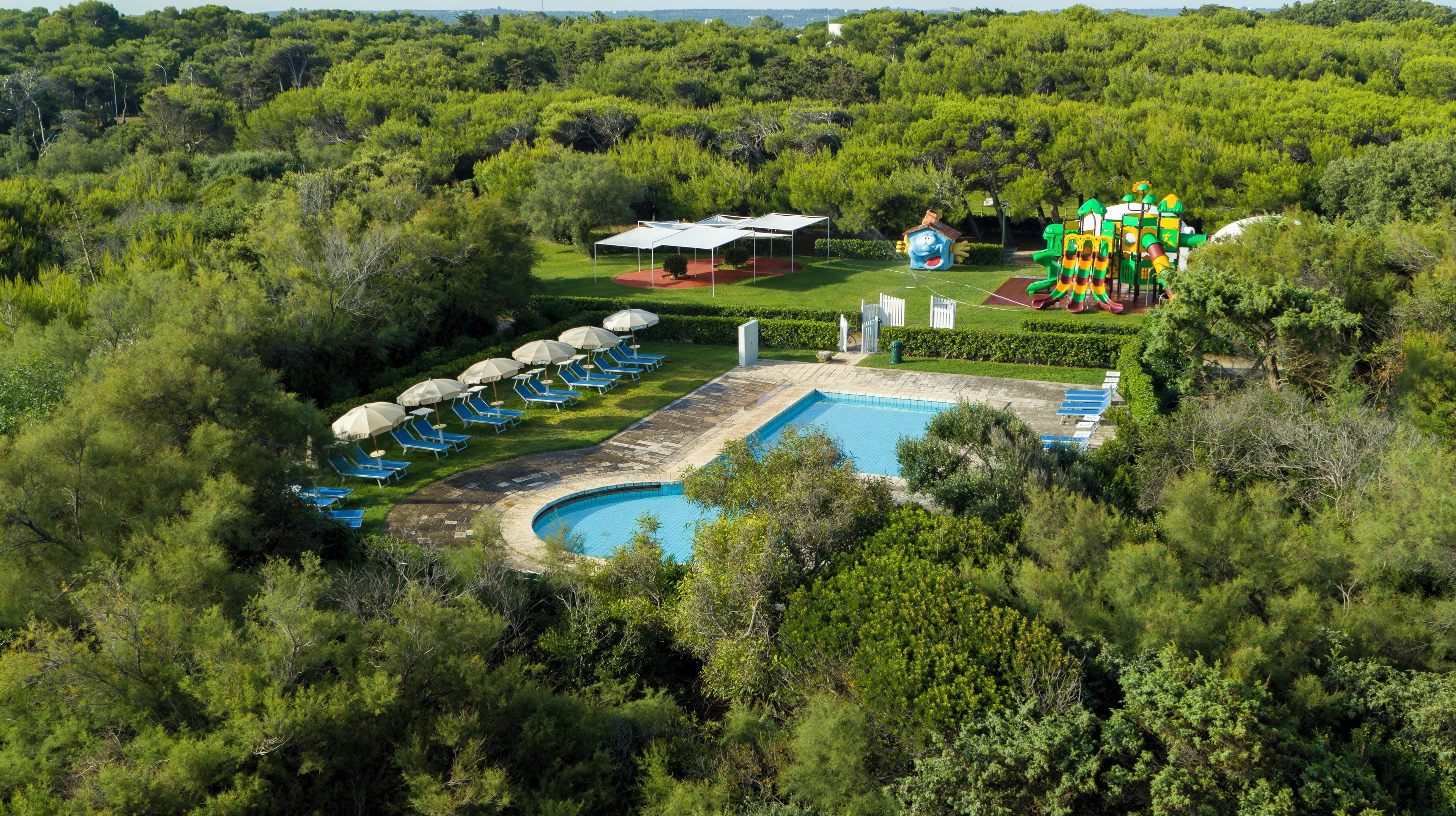 Una vista aerea di un resort con piscina, lettini prendisole e un’area giochi per bambini.