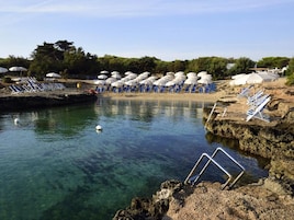 A beach with clear water, sun loungers, umbrellas, and a small pool area.