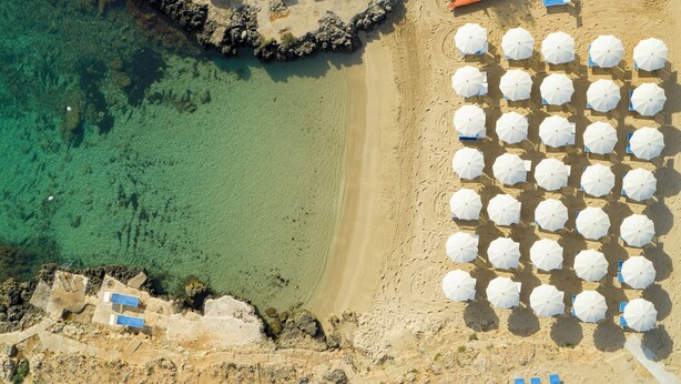 Vista aérea de una playa con agua turquesa cristalina, una orilla de arena y numerosas sombrillas blancas dispuestas en filas.