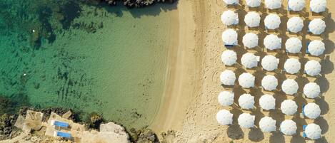 An aerial view of a beach with clear turquoise water, a sandy shore, and multiple white umbrellas arranged in rows.