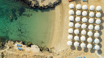 An aerial view of a beach with clear turquoise water, a sandy shore, and multiple white umbrellas arranged in rows.