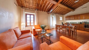 Un amplio salón con techo de madera, sofás naranja, una mesa de centro de cristal y un comedor con sillas de madera. Se observa una cocina al fondo.
