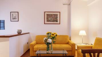A living room with a yellow sofa, a glass coffee table, and a vase with flowers. There are two framed pictures on the wall and a lamp on a side table.
