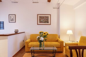 A living room with a yellow sofa, a glass coffee table, and a vase with flowers. There are two framed pictures on the wall and a lamp on a side table. - Tenuta Montecucco (Cinigiano)