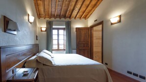 Un dormitorio con techo de madera, una cama con ropa de cama blanca, una mesita de noche, una ventana con cortinas y un armario de madera.