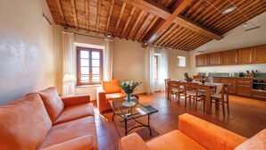 A spacious living area with a wooden ceiling, orange sofas, a glass coffee table, and a dining area with wooden chairs and a kitchen in the background. - Tenuta Montecucco (Cinigiano)