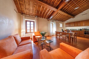 A spacious living area with a wooden ceiling, orange sofas, a glass coffee table, and a dining area with wooden chairs and a kitchen in the background. - Tenuta Montecucco (Cinigiano)