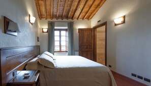 A bedroom with a wooden ceiling, a bed with white linens, a nightstand, a window with curtains, and a wooden wardrobe. - Tenuta Montecucco (Cinigiano)