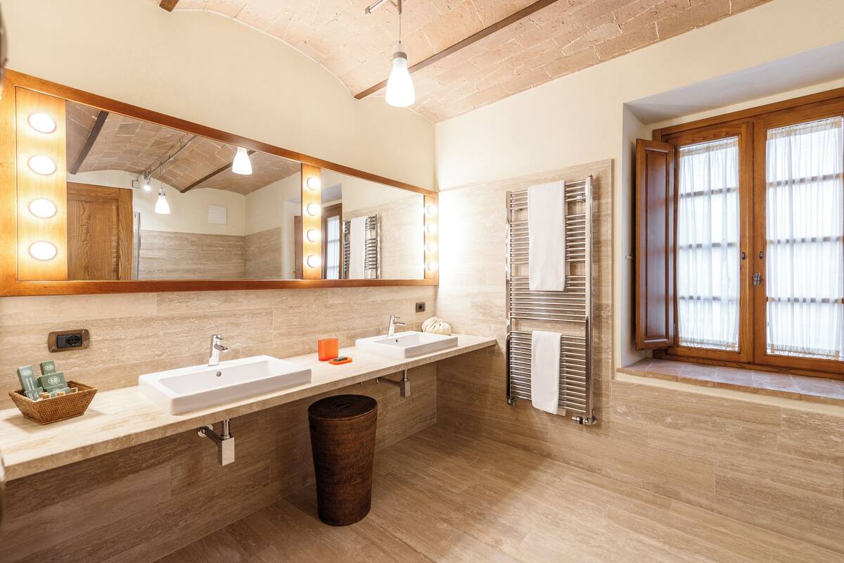 A modern bathroom with two sinks, a large mirror with lights, a wooden ceiling, and a window with curtains.