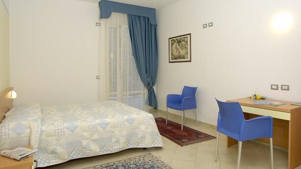 A bedroom with a bed, a desk, two chairs, a window with curtains, and a wall-mounted light fixture. - Residence Rosmari (Brenzone sul Garda)