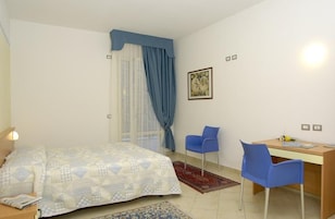Un dormitorio con cama, escritorio, dos sillas, ventana con cortinas e iluminación en la pared.