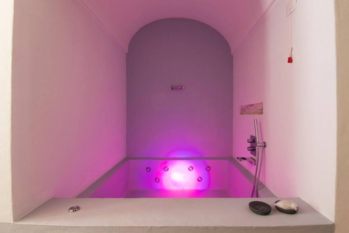 Un baño moderno con una bañera iluminada en morado, ducha y un estante pequeño con objetos.