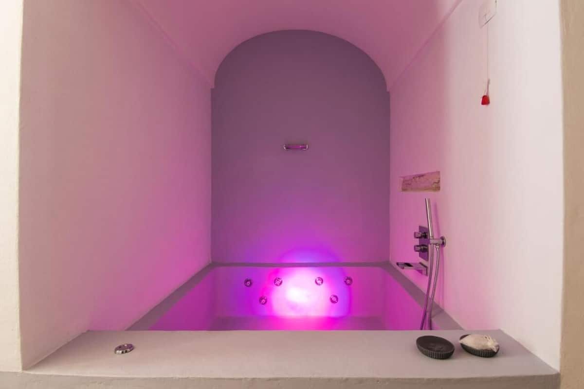 Une salle de bain moderne avec une baignoire éclairée en rose, une douche et une petite étagère avec quelques objets.