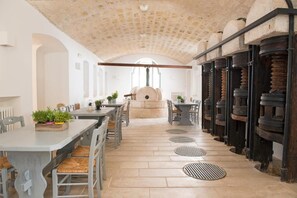 A spacious dining area with a long table and chairs, a stone ceiling, and a view of the kitchen area. - Masseria San Giovanni (Fasano)