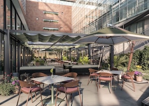 An outdoor dining area with tables and chairs, surrounded by plants and a modern building in the background. - Hyatt Regency Rome Central (Rome)