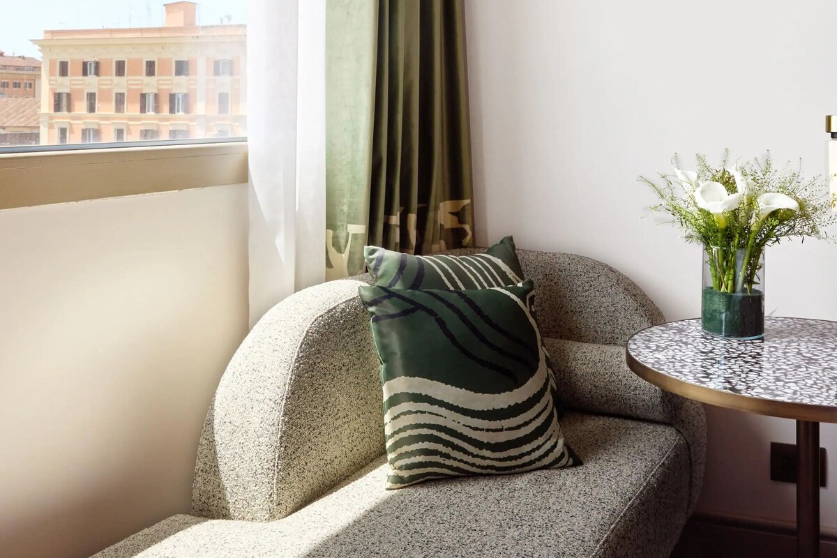 A corner seating area with a textured sofa, a patterned pillow, a small round table with a glass vase of flowers, and a view of buildings outside.