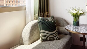 A corner seating area with a textured sofa, a patterned pillow, a small round table with a glass vase of flowers, and a view of buildings outside.