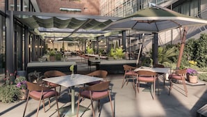 An outdoor dining area with tables and chairs, surrounded by plants and a modern building in the background. - Hyatt Regency Rome Central (Rome)