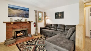 A living room with a brick fireplace, a gray sofa, and a large painting of a snowy landscape. - Sugar Bear Chalet #2559 (Sugarloaf)