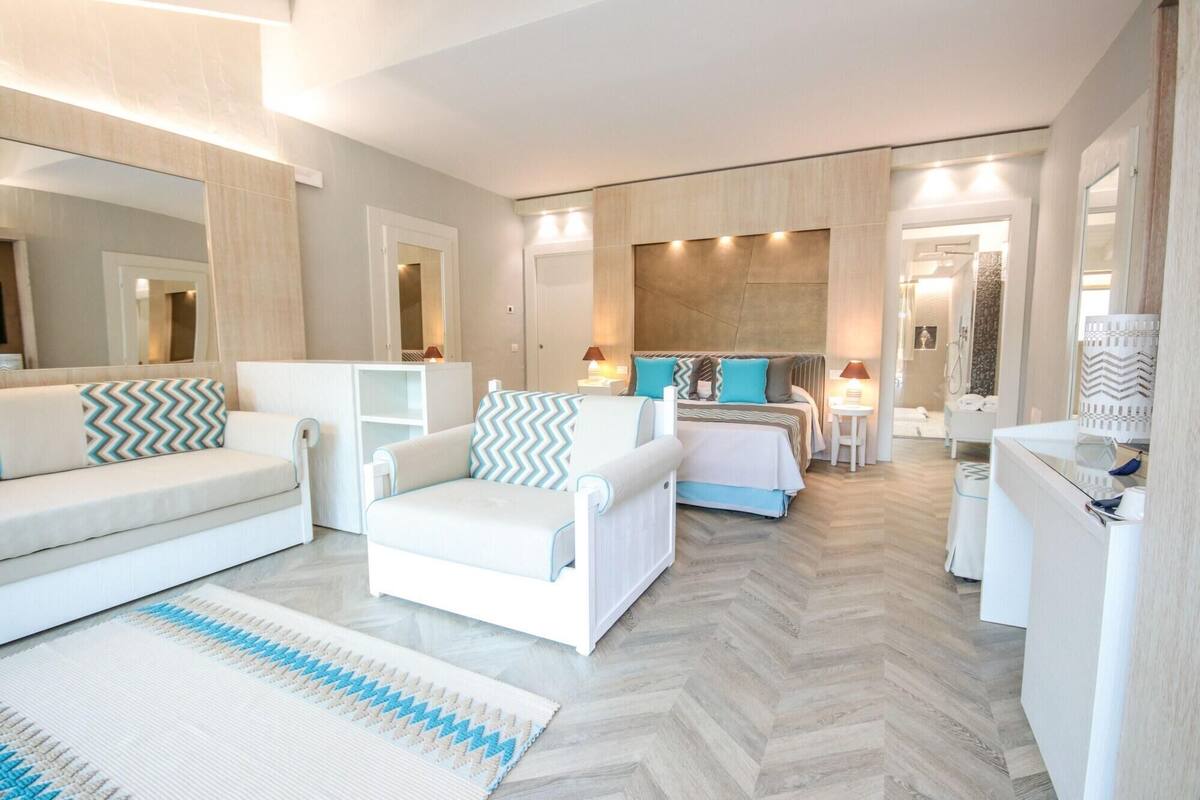 A modern hotel room with a sofa, armchair, and a bed with patterned pillows. There is a small table with a lamp and a mirror on the wall.