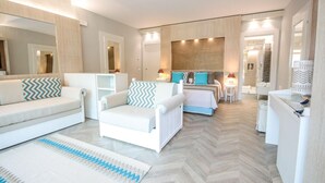 A modern hotel room with a sofa, armchair, and a bed with patterned pillows. There is a small table with a lamp and a mirror on the wall. - Eliantos Hotel and Spa (Pula)