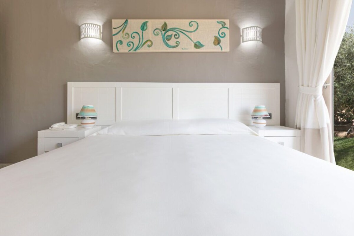 A bedroom with a white bed, two bedside lamps, a decorative wall piece, and a view of greenery outside.