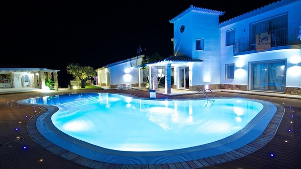 A swimming pool with a wooden deck surrounding it, a building with a balcony, and a well-lit entrance. - Eliantos Hotel and Spa (Pula)