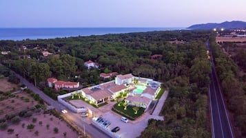 An aerial view of a residential area with a large house featuring a swimming pool, surrounded by dense greenery and a road.