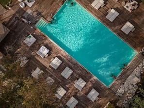 Eine Vogelperspektive auf einen rechteckigen Swimmingpool, umgeben von einer Holzterrasse mit Liegestühlen und einem kleinen Wasserfall.