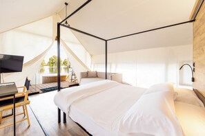 Ein modernes Schlafzimmer mit einem großen Bett, einem Schreibtisch und einem Fernseher. Ein Balkon bietet Blick auf Bäume und ein Gebäude.