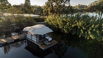 Una casa flotante con terraza techada y sillas de descanso, rodeada de agua y árboles.