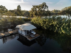Una casa flotante con terraza techada y sillas de descanso, rodeada de agua y árboles.