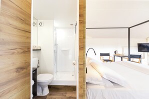 Un baño moderno con ducha, inodoro y lavamanos. La habitación tiene suelo de madera y una cama con televisor y sillas.