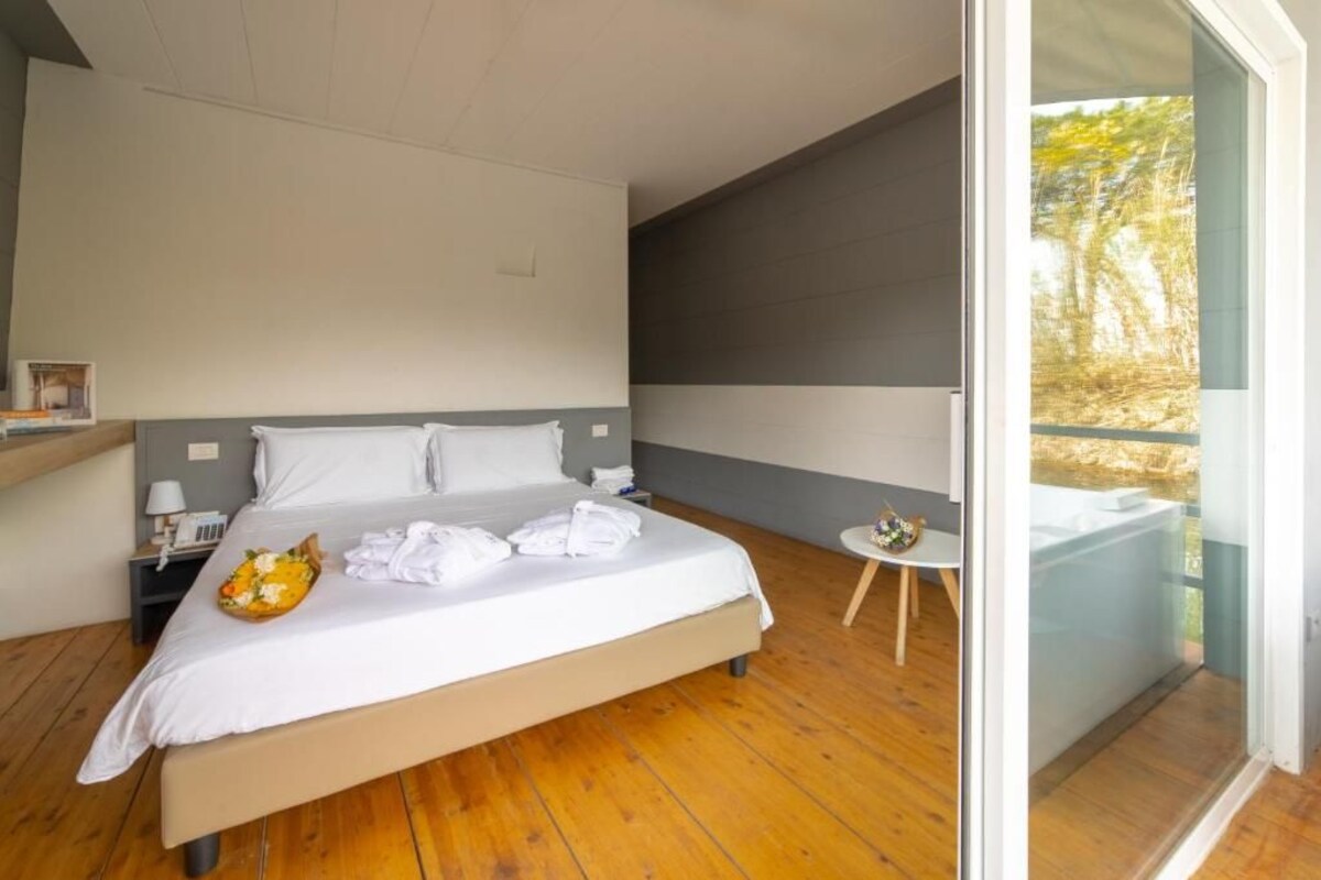 A modern bedroom with a large bed, a bedside table with a lamp, and a view of a balcony with a hot tub.